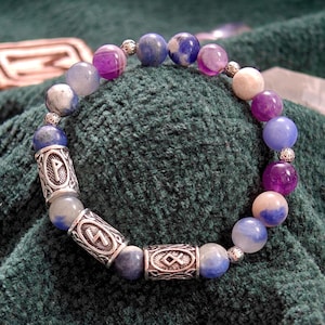 Op de afbeelding: Een kralenarmband met paarse, blauwe en witte edelstenen. De armband heeft zilverkleurige cilindrische kralen met geëtste ontwerpen. De kralen zijn geregen op een zilverkleurige ketting. De armband wordt getoond op een donkergroene stof.