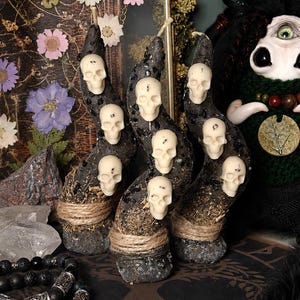 May include: Three curved, dark-textured candles adorned with multiple small, off-white skull figures. Each candle base is wrapped in natural twine and features a speckled, stone-like finish. The candles are arranged upright in a cluster.