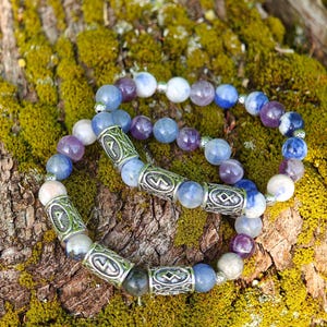 May include: Two beaded bracelets with blue, purple, and white stones. The bracelets have silver-colored cylindrical beads with etched designs. The bracelets are resting on a mossy, textured surface.