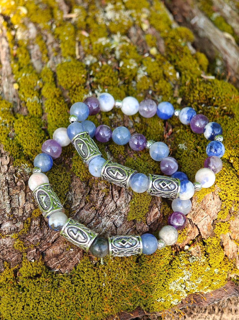 May include: Two beaded bracelets with blue, purple, and white stones. The bracelets have silver-colored cylindrical beads with etched designs. The bracelets are resting on a mossy, textured surface.