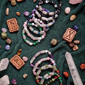 May include: An assortment of gemstone bracelets in various colors, including green, pink, purple, and black, with silver-colored metal accents. The bracelets are displayed with wooden rune tiles, crystals, and polished stones on a dark green fabric.