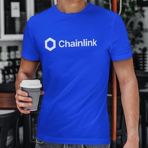May include: Royal blue t-shirt with the white Chainlink logo and text. The logo is a hexagon shape. The shirt is short-sleeved and made of a soft material. A person is holding a gray coffee cup.