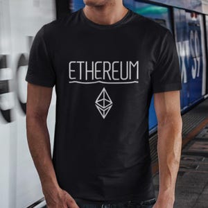 Puede incluir: Camiseta negra con la palabra "ETHEREUM" en blanco, sobre un logotipo blanco de Ethereum. La camiseta de manga corta parece ser de algodón o un material similar. El fondo es una vista borrosa de una estación de tren.