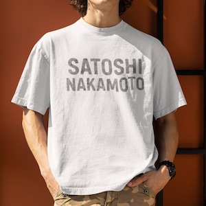 May include: A white t-shirt with the name "SATOSHI NAKAMOTO" printed in gray, distressed lettering. The shirt is a crew neck style and appears to be made of a soft, comfortable material. The person is wearing a black watch and khaki pants.