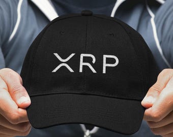 May include: Black baseball cap with the white letters "XRP" across the front. The cap is held up by a person. The hat has a curved brim and a rounded top.