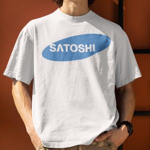May include: White t-shirt with a blue oval design that reads "SATOSHI" in white capital letters. The shirt is short-sleeved and appears to be made of cotton. The person wearing the shirt has a black watch on their wrist.