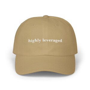 May include: A tan baseball cap with the phrase "highly leveraged" embroidered in white across the front. The cap has a curved brim and a rounded crown. The hat is made of a soft, durable fabric.