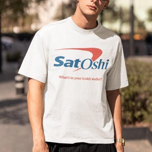 May include: A light gray t-shirt with the word "Satoshi" in blue and red, and the phrase "What's in your (cold) wallet?" in red. The shirt is a crew neck style.