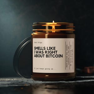 May include: A brown glass jar candle with a lit wick. The label reads "SMELLS LIKE I WAS RIGHT ABOUT BITCOIN." The candle is hand-poured in the USA and has a 50-hour burn time. The jar is open, revealing the lid.