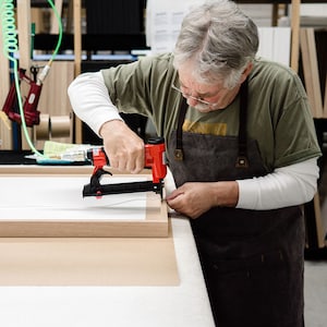 May include: A person using a red electric staple gun to attach a wooden frame to a white canvas. The person is wearing a green t-shirt and a brown apron.