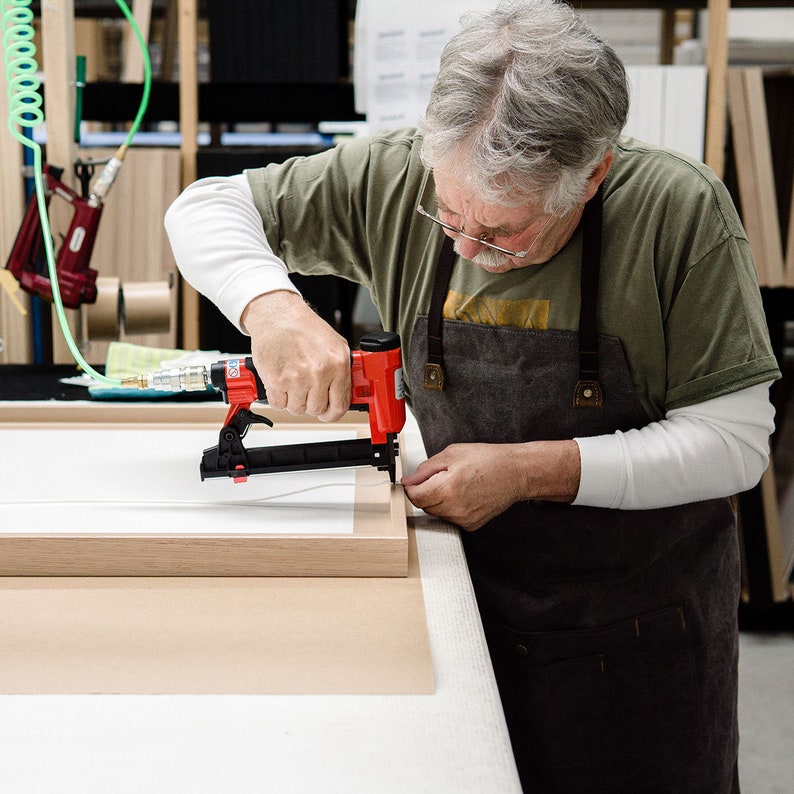 May include: A person using a red electric staple gun to attach a wooden frame to a white canvas. The person is wearing a green t-shirt and a brown apron.