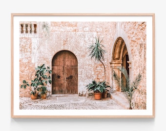 May include: A framed print of a European courtyard scene. The image features a weathered stone wall with a wooden arched doorway and a wooden door. Potted plants and a cobblestone path add to the rustic charm. The print is in a light wood frame.