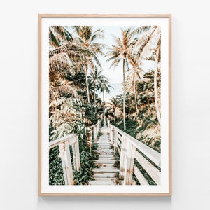 May include: A wooden walkway with railings leads through a lush tropical forest with palm trees and green foliage. The path leads to a beach with blue water in the distance.