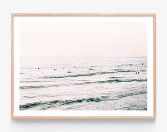 May include: A photograph of a seascape with surfers in the distance. The water is a light blue and green color, and the sky is a soft pink. The image is framed in a light wood frame.