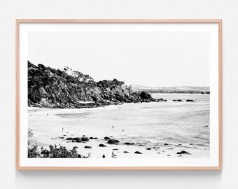 May include: Black and white photograph of a rocky coastline with a sandy beach and ocean waves. There are people in the distance on the beach.