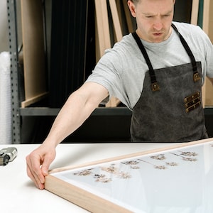 May include: A person in a grey t-shirt and a brown apron is framing a print of a landscape with tall, thin trees. The print is in a light wood frame.