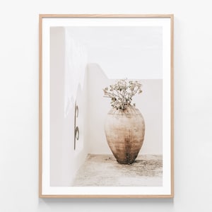 May include: A framed print showing a large, aged terracotta vase with greenery. The vase is set against a white wall and a light-coloured floor, creating a minimalist, neutral aesthetic. The artwork is framed in light wood.