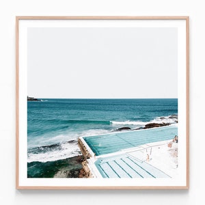 May include: An aerial view of an outdoor swimming pool with blue water, surrounded by a white concrete deck. The pool is located on a rocky coastline with crashing waves in the background.