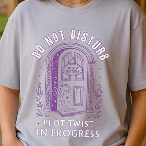 May include: A light gray t-shirt with a purple graphic that reads "DO NOT DISTURB" above a door design and "PLOT TWIST IN PROGRESS" below. The design includes candles, a book, and a butterfly.