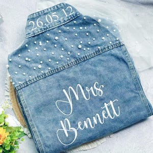 Op de afbeelding: Lichtblauwe spijkerjas versierd met witte parels. De jas heeft de datum "26.05.12" en de naam "Mrs. Bennett" in witte letters. Een decoratief item voor een bruiloft of bruidsevenement.