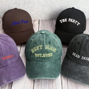 May include: Five baseball caps in different colours, each embroidered with a unique phrase. The caps are brown, black, green, purple, and grey. The phrases include "Boat Daddy," "Best Man Est. 2025," and "Dead Inside."