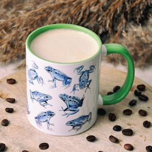 Può includere: Una tazza di ceramica bianca con manico e bordo verdi, piena di una bevanda cremosa. La tazza è decorata con illustrazioni di rane blu. Chicchi di caffè sparsi intorno alla tazza su una superficie di legno.