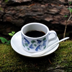 Puede incluir: Un juego de taza y platillo de café de cerámica blanca con un diseño de rana y nenúfar, lleno de café oscuro. Una cuchara blanca descansa sobre el platillo. El juego se encuentra sobre un tronco cubierto de musgo, con un fondo borroso de bosque.