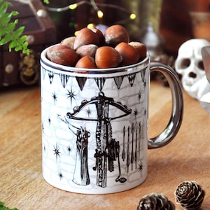 May include: A white ceramic mug featuring a black ink illustration of a crossbow, a quiver of arrows, and a banner of triangular flags against a brick wall background. The mug is filled with a collection of whole brown hazelnuts.