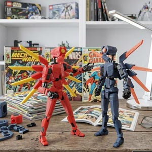 May include: Two articulated robot action figures, one red with yellow accents and the other dark blue with orange accents, stand on a wooden desk. Comic books and building blocks are scattered around. The figures have wing-like structures and are posed as if in action.