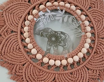 Könnte beinhalten: Staubrosa Makramee-Wandbehang mit kreisförmigem Design. Die Mitte zeigt eine Glasscheibe mit einem geätzten Elefanten und floralen Details. Holzperlen sind um den inneren Kreis herum angeordnet und verleihen dieser Boho-Dekoration Textur.