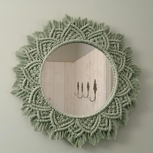 Puede incluir: Espejo redondo de macramé verde salvia. El marco presenta un diseño detallado en forma de sol con un borde con flecos. El espejo refleja una pared de madera clara con ganchos decorativos. El diámetro del espejo es de unos 40 cm.