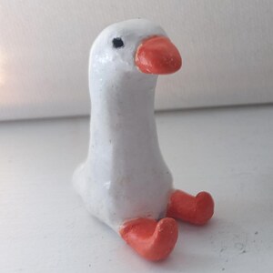 Op de afbeelding: Een handgemaakt wit ganzenbeeldje met een feloranje snavel en poten. De gans zit, met de poten uitgestrekt. Het beeldje heeft een gladde, geschilderde afwerking en een klein zwart oog.