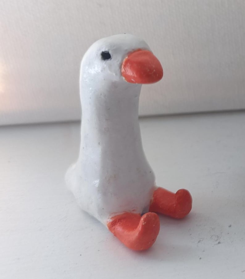 Op de afbeelding: Een handgemaakt wit ganzenbeeldje met een feloranje snavel en poten. De gans zit, met de poten uitgestrekt. Het beeldje heeft een gladde, geschilderde afwerking en een klein zwart oog.