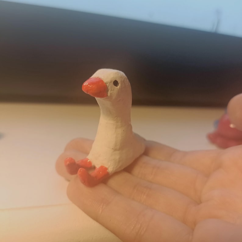Op de afbeelding: Een kleine, handgemaakte ganzenfiguur. De gans is wit met een felrode snavel en poten. Hij zit met uitgestrekte poten. Het beeldje rust op de open handpalm van een persoon.