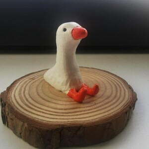 Op de afbeelding: Handgemaakte witte ganzenfiguur met een oranje snavel en poten, geplaatst op een ronde houten schijf. De gans zit met zijn kop omhoog. De houten schijf heeft zichtbare groeiringen en een donkerbruine schorsrand.