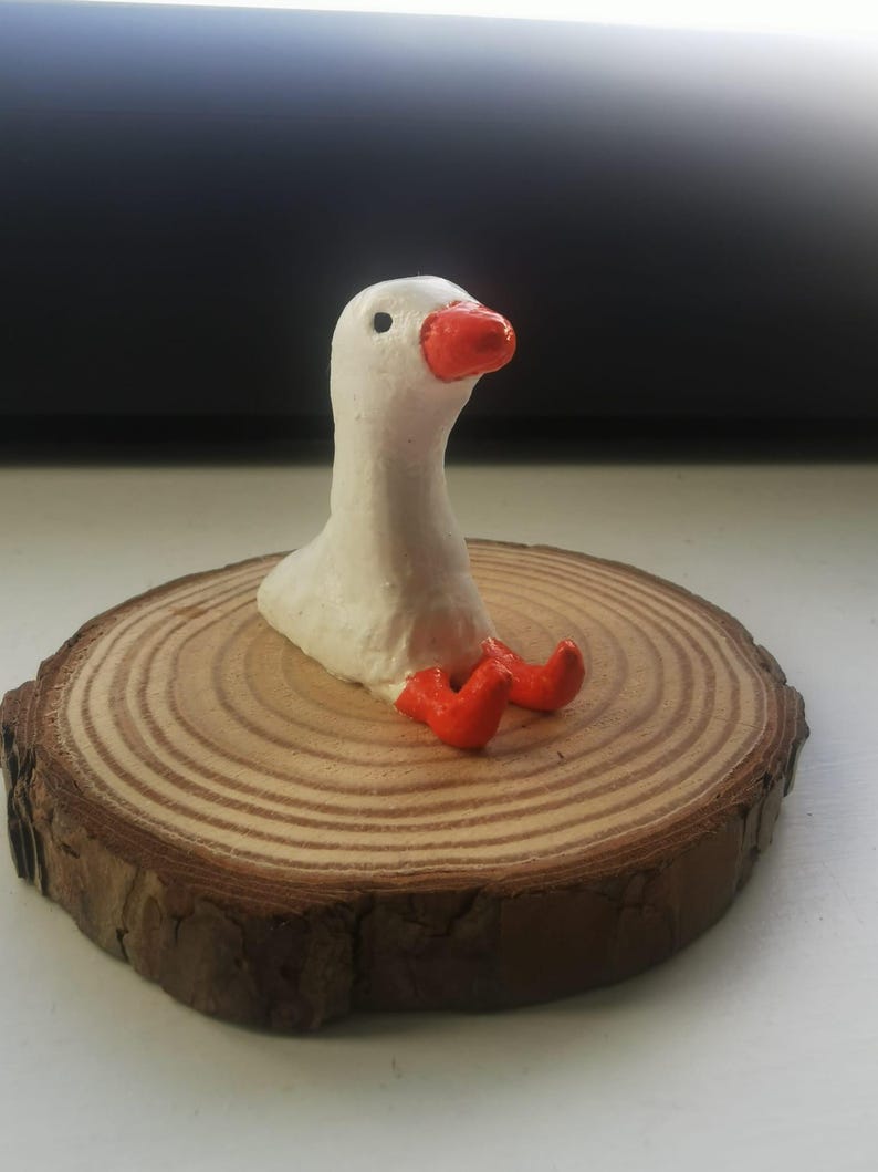 Op de afbeelding: Handgemaakte witte ganzenfiguur met een oranje snavel en poten, geplaatst op een ronde houten schijf. De gans zit met zijn kop omhoog. De houten schijf heeft zichtbare groeiringen en een donkerbruine schorsrand.
