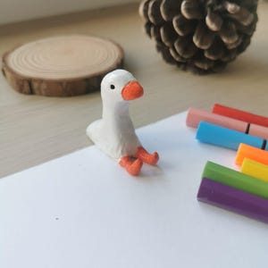 Op de afbeelding: Een kleine, witte ganzenfiguur met een oranje snavel en poten zit op een wit oppervlak. Een houten schijf en een dennenappel zijn op de achtergrond te zien. Gekleurde stiften suggereren een knutsel- of kunstthema.