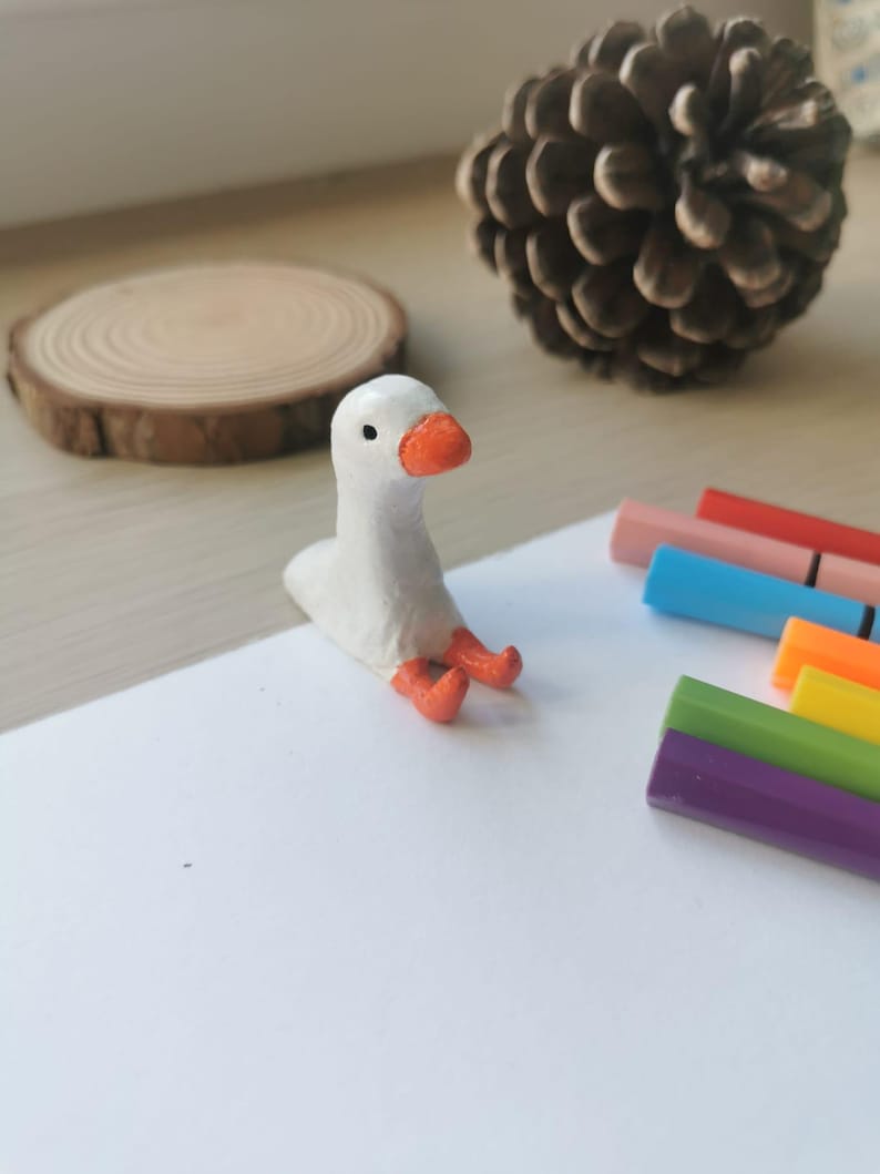 Op de afbeelding: Een kleine, witte ganzenfiguur met een oranje snavel en poten zit op een wit oppervlak. Een houten schijf en een dennenappel zijn op de achtergrond te zien. Gekleurde stiften suggereren een knutsel- of kunstthema.