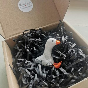 Op de afbeelding: Een kleine, witte ganzenfiguur met een oranje snavel en poten rust in een kartonnen doos gevuld met zwarte papiersnippers. De doos heeft een sticker met de tekst "thank you for your order".
