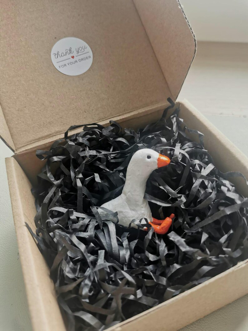 Op de afbeelding: Een kleine, witte ganzenfiguur met een oranje snavel en poten rust in een kartonnen doos gevuld met zwarte papiersnippers. De doos heeft een sticker met de tekst "thank you for your order".