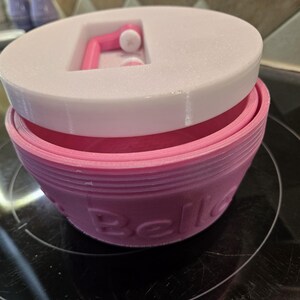 May include: A two-tone container with a pink base and a white lid. The pink base has the word "Belle" embossed on the side. The lid has a recessed area with a pink handle. The container is sitting on a black surface.