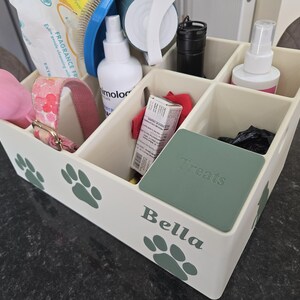 May include: A cream-colored plastic storage organizer with six compartments, decorated with green paw prints and the name Bella. The box contains various pet care items, including a pink collar, a spray bottle, and a green lid labeled Treats.
