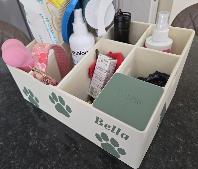 May include: A cream-colored plastic storage organizer with six compartments, decorated with green paw prints and the name Bella. The box contains various pet care items, including a pink collar, a spray bottle, and a green lid labeled Treats.