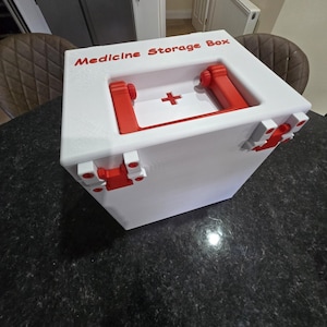 Puede incluir: Una caja de almacenamiento de medicamentos rectangular de color blanco con detalles en rojo. La parte superior presenta un asa roja, un símbolo de cruz roja y el texto Medicine Storage Box impreso en rojo. Incluye bisagras y cierres rojos laterales.