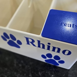 May include: A white plastic storage container with multiple compartments, featuring blue paw print graphics and the name Rhino printed in blue. A blue lid with the embossed text Treats sits on top of one of the compartments of the organizer.