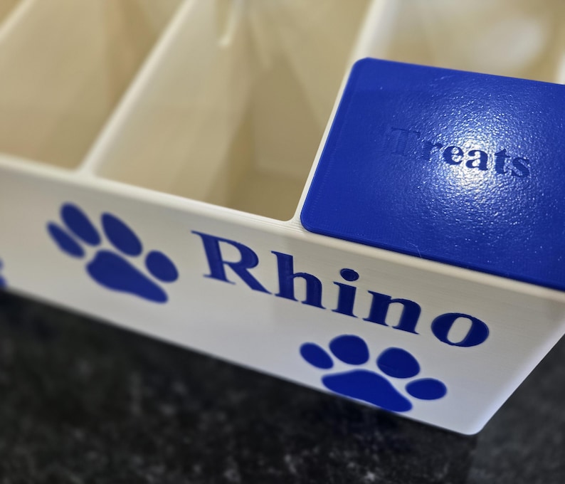 May include: A white plastic storage container with multiple compartments, featuring blue paw print graphics and the name Rhino printed in blue. A blue lid with the embossed text Treats sits on top of one of the compartments of the organizer.