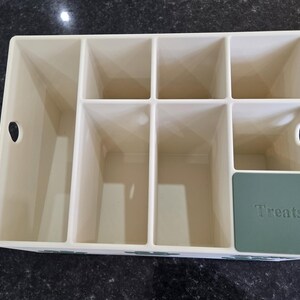 May include: A cream-colored plastic storage organizer with multiple rectangular compartments of varying sizes. The unit features two circular side cutouts for handles and a dark green square panel on the front right corner with the word Treats embossed.