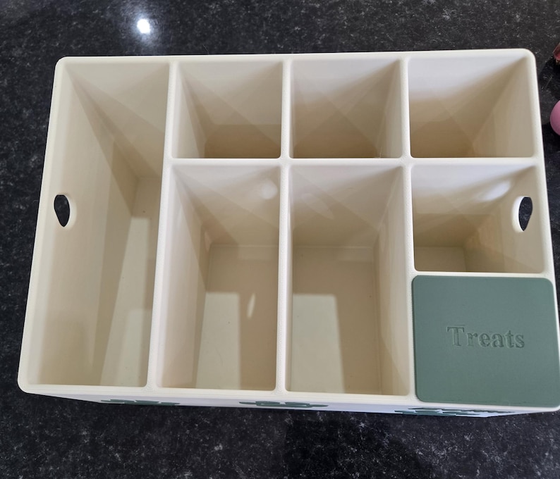 May include: A cream-colored plastic storage organizer with multiple rectangular compartments of varying sizes. The unit features two circular side cutouts for handles and a dark green square panel on the front right corner with the word Treats embossed.