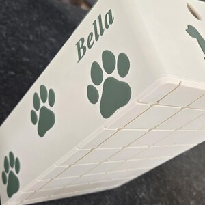 May include: A cream-colored rectangular container featuring the name Bella in dark green script lettering, accompanied by several dark green paw print graphics. The side of the container displays a textured grid pattern of small squares.