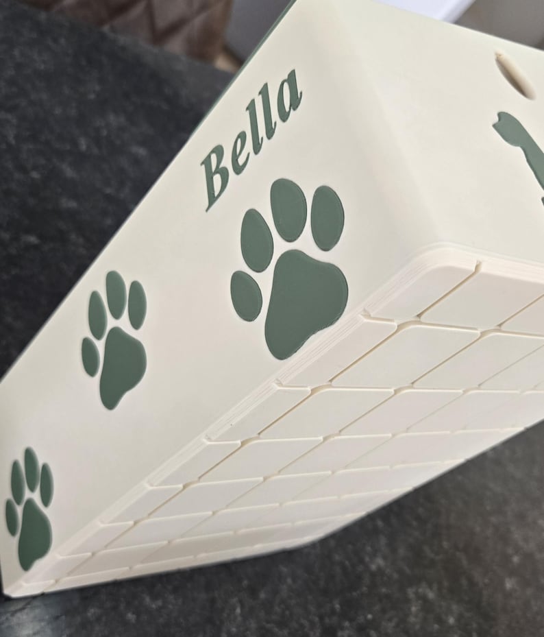 May include: A cream-colored rectangular container featuring the name Bella in dark green script lettering, accompanied by several dark green paw print graphics. The side of the container displays a textured grid pattern of small squares.